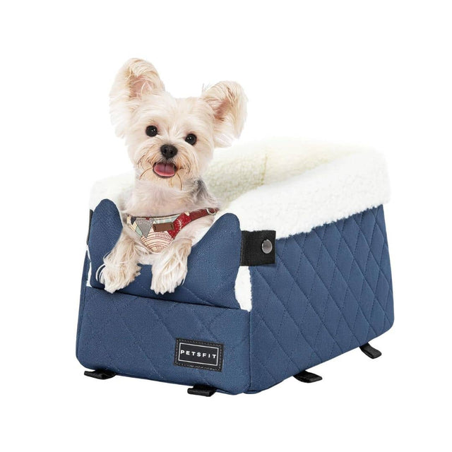 Little dog sits upright in a blue quilted dog car seat with plush white interior, posed against a light backdrop.