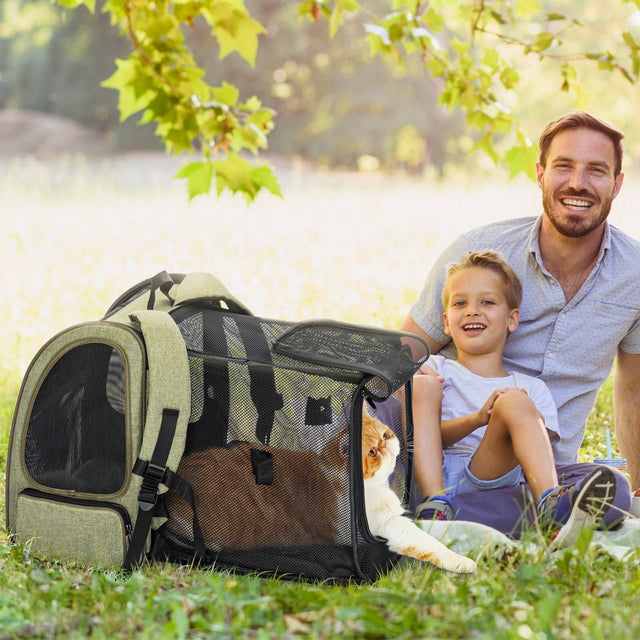 Outdoor picnic scene with father and child beside expanded cat backpack; mesh sides open as cat lounges inside on grass.