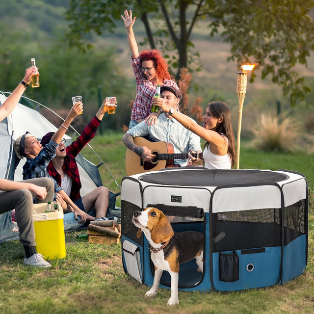 Portable playpen set on grass at a campsite while friends relax nearby, keeping a beagle safely contained outdoors.
