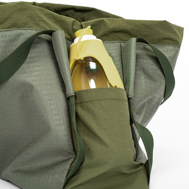 Olive gear bag for dog backpack trips, carried by hand with long handles, side pockets, and tan work boots.