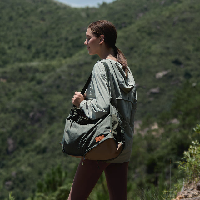 Olive gear bag for dog backpack trips, carried by hand with long handles, side pockets, and tan work boots.