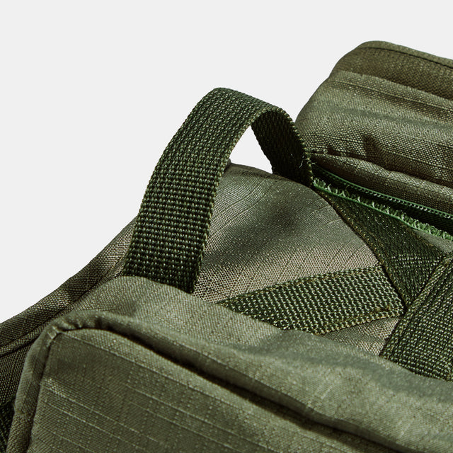 Detailed view of the top grab handle and crisscross webbing stitched onto olive fabric beside the upper storage pocket zipper.