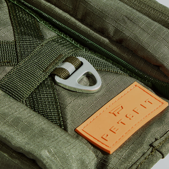 Macro shot of silver triangular leash ring and embossed logo patch on olive fabric, with zipper edge and webbing attachment visible.
