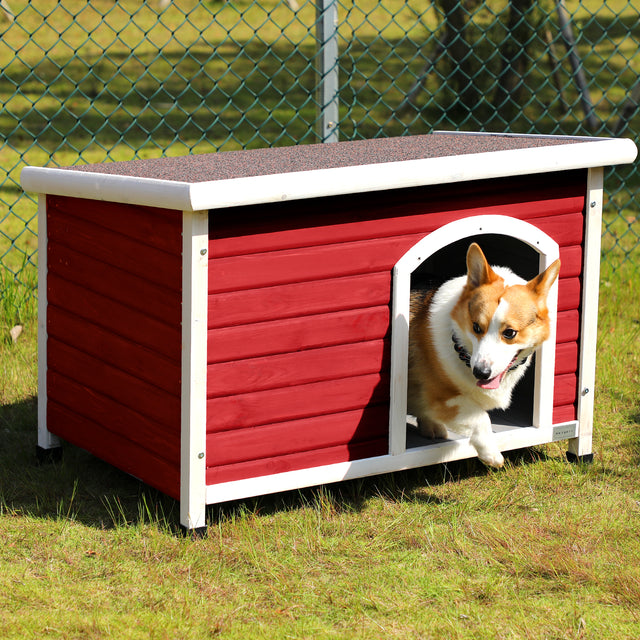 Corgi steps out of a red outdoor dog house beside a chain-link fence, showing a roomy entrance and raised base on grass.