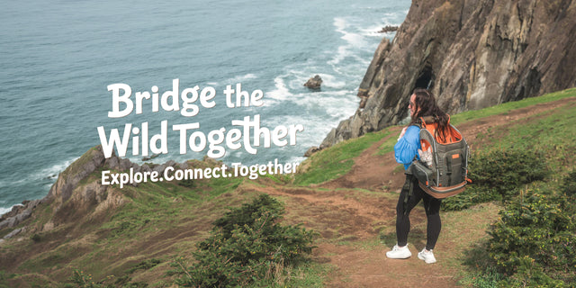 Hiker on a coastal cliff trail with a tabby cat riding in an orange-lined grey cat backpack above the breaking surf