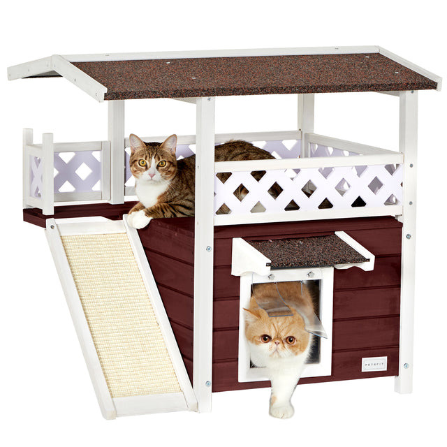 Two cats use a brown-and-white outdoor cat house, one resting on the balcony and one stepping through the curtained front door.