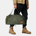 Olive gear bag for dog backpack trips, carried by hand with long handles, side pockets, and tan work boots.