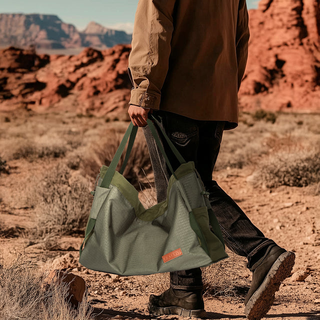 Olive gear bag for dog backpack trips, carried by hand with long handles, side pockets, and tan work boots.