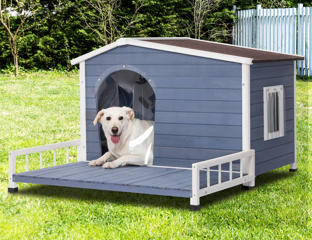 White dog lounges just inside the clear flap entrance of a raised outdoor dog house set on a bright green lawn.